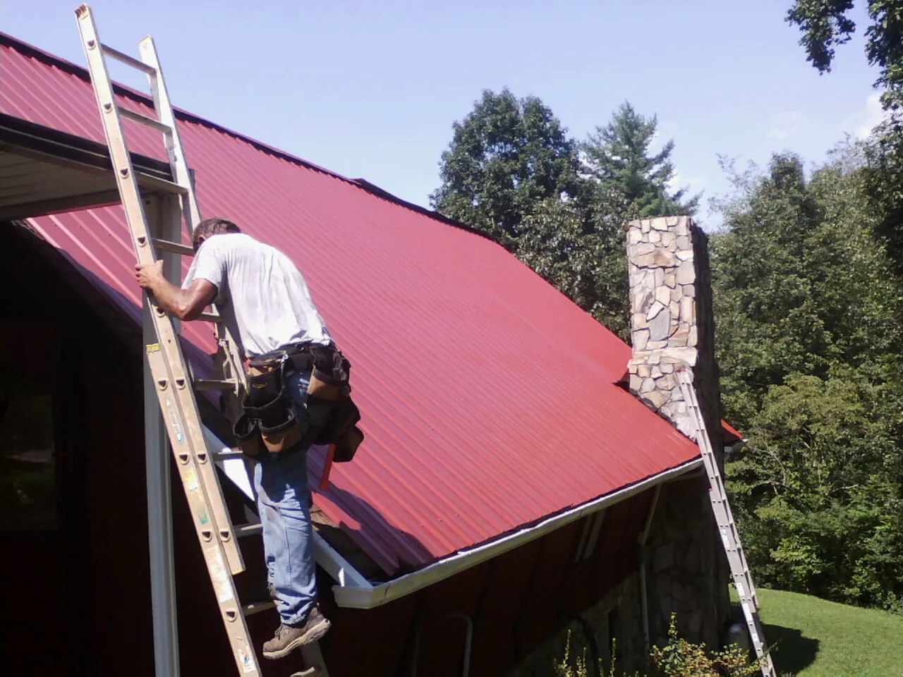 Roofer on a ladder beside a red metal roof and stone chimney — Soffit & Fascia Repair by Roofing Brothers in Kenmore